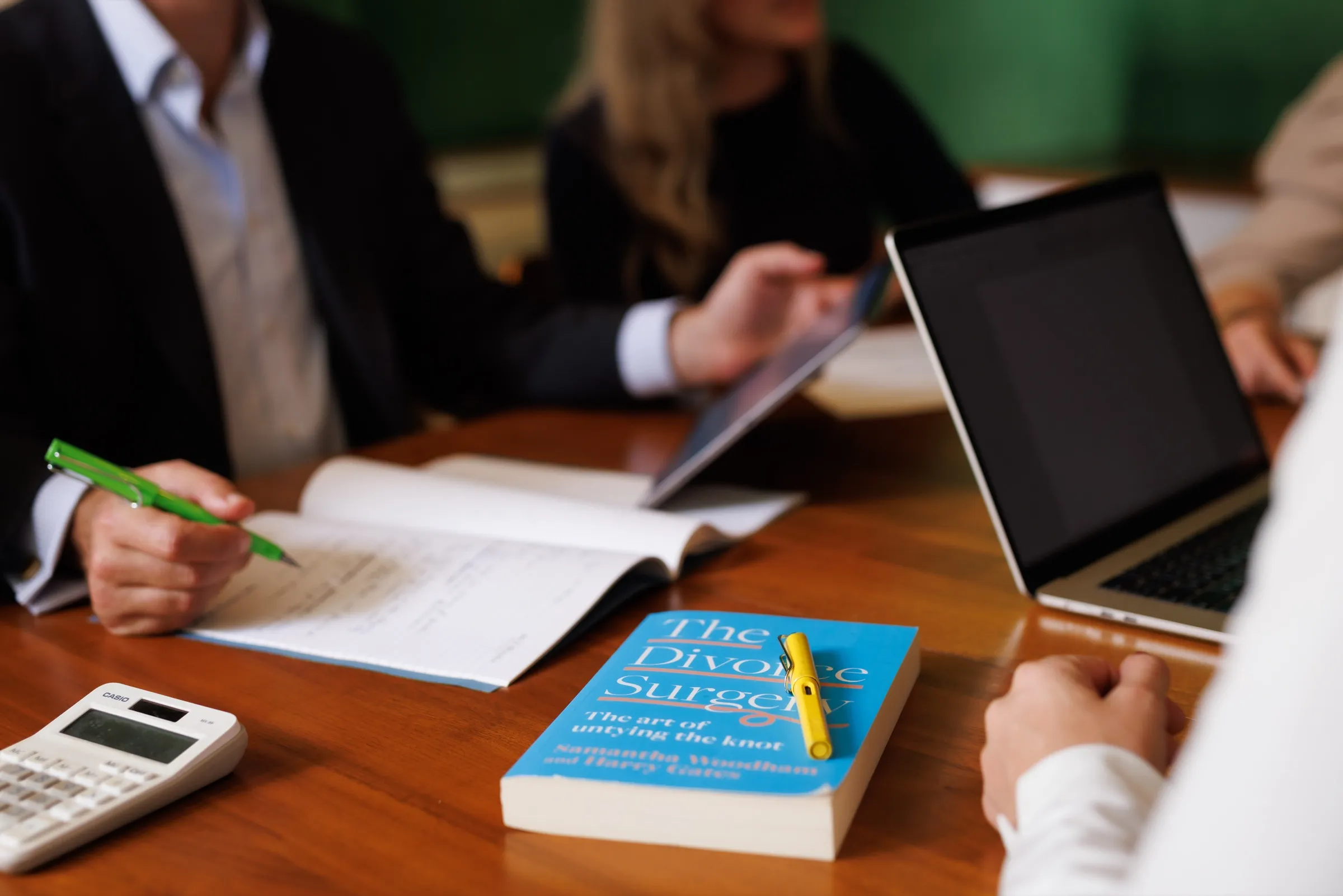 The Divorce Surgery book on a meeting table beside a notebook, calculator and laptop, with the team working in the background