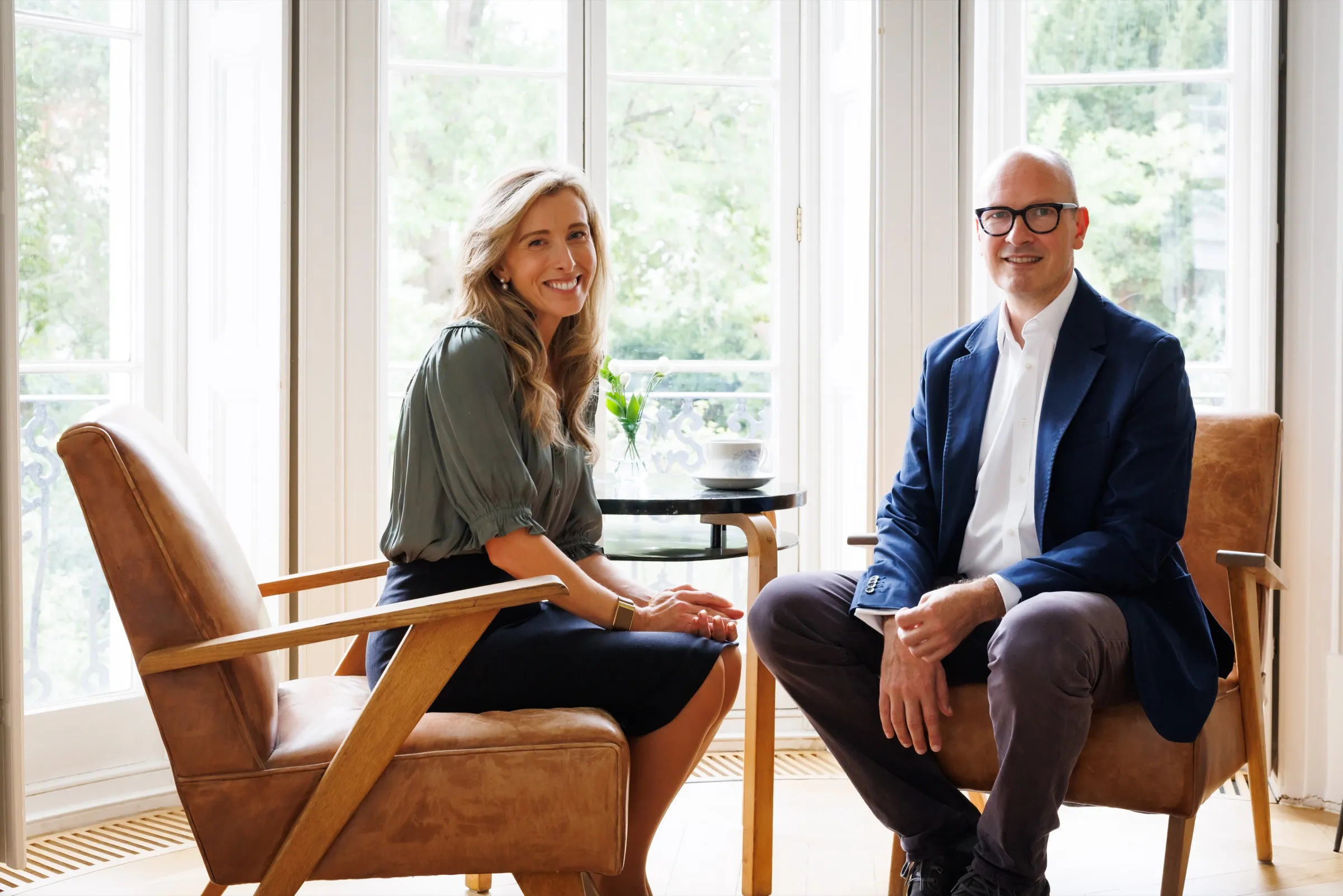 Two people seated in leather armchairs by a sunlit window, smiling towards the camera