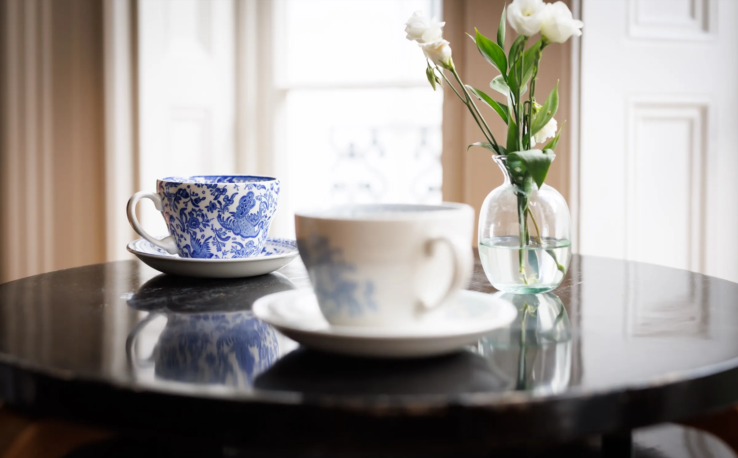 Two teacups and a vase of white flowers on a marble table by a window